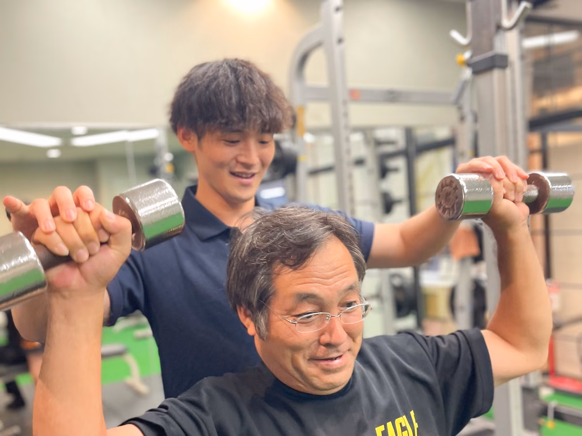 ダンベルを両手で持ち上げる会員の後ろで、トレーナーが両腕を支えている様子。