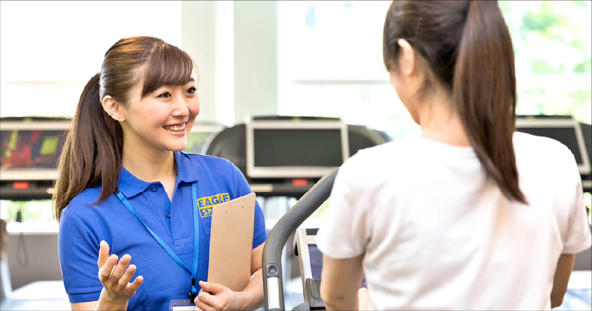 フィットネスジムでクリップボードを持ち笑顔で案内するトレーナー