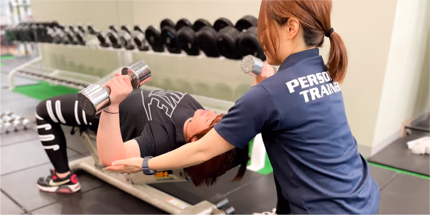 パーソナルトレーナーがベンチプレスのフォームをサポートしながら女性にダンベルを使ったトレーニングを指導しているジムの様子。