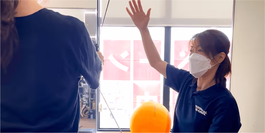 マスクを着用した女性トレーナーが手を挙げて、別の人物に何かを示している室内のシーン。