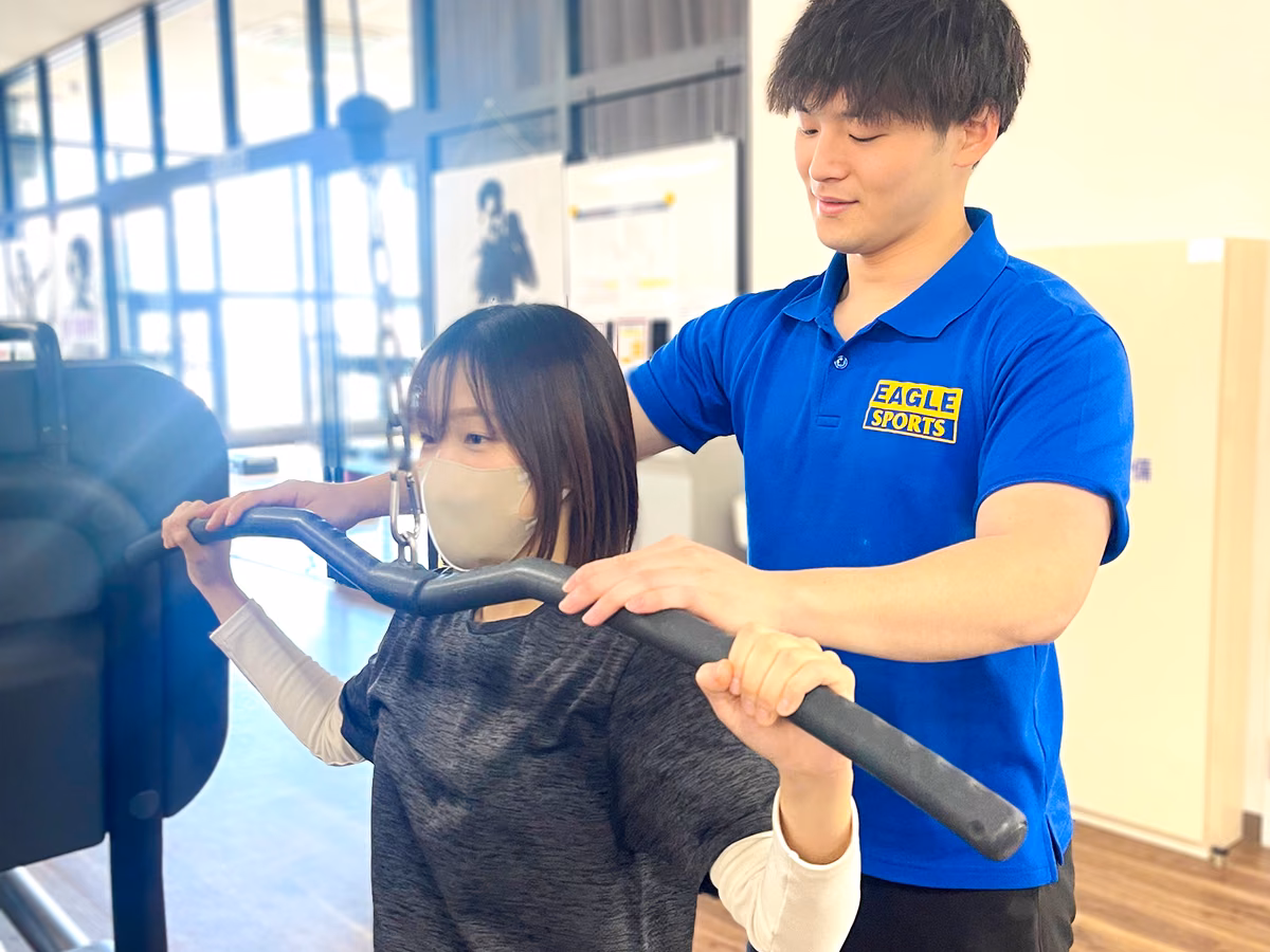 マシンのバーを握る会員の後ろで、トレーナーが姿勢を支えている様子。