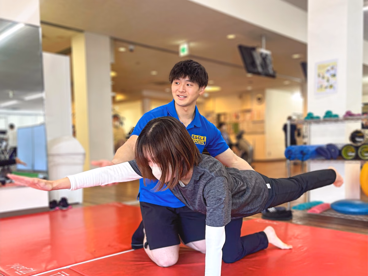 赤いマットの上で片脚と片腕を伸ばす会員を、後ろでトレーナーが支えている様子。