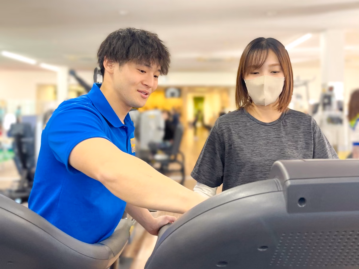ランニングマシンの操作パネルを、会員の横でトレーナーが指し示している様子。