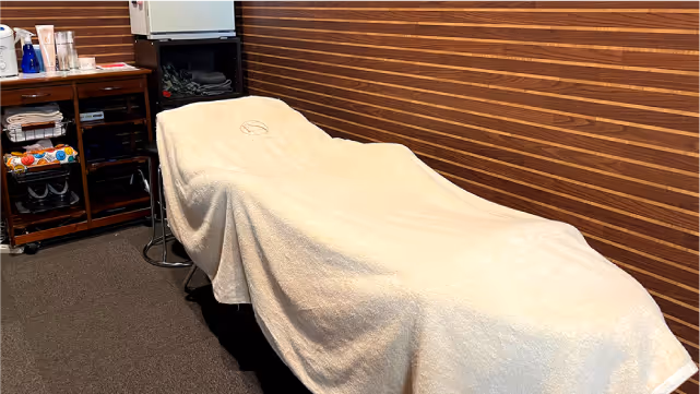 ベージュのタオルがかけられたマッサージベッドが、木のストライプ壁のある部屋に置かれている。