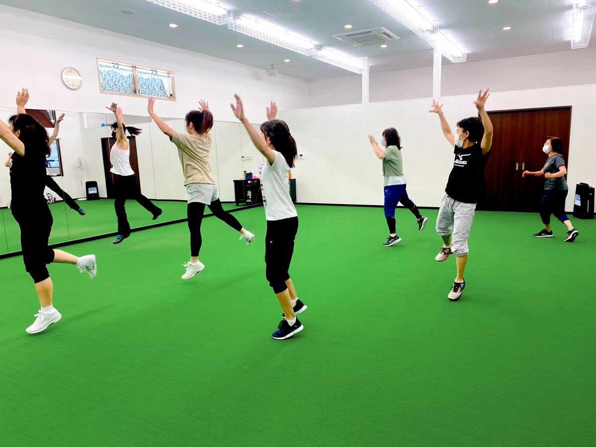 緑のフロアのスタジオで、参加者が両手を上げて跳びながら動いているレッスンの様子。