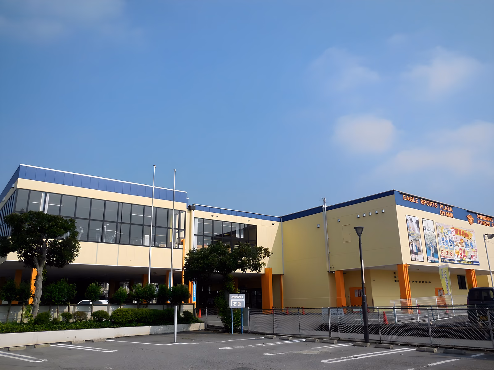 青空の下、屋根に「EAGLE SPORTS PLAZA OYAMA」の看板を掲げたクリーム色と紺色の建物。手前に広い駐車場が広がる。