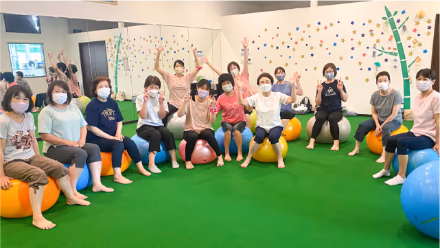 マスクを着用した女性たちがカラフルなバランスボールの上に座り、緑の床の上で手を挙げている室内グループクラス。