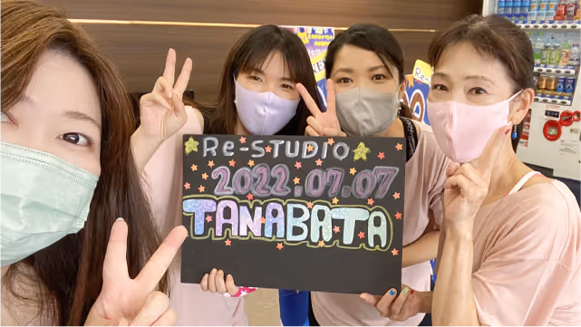 マスクを着けた4人の女性が、手でピースを作りながら「2022 07 07 TANABATA」と書かれた黒いボードを持っている。